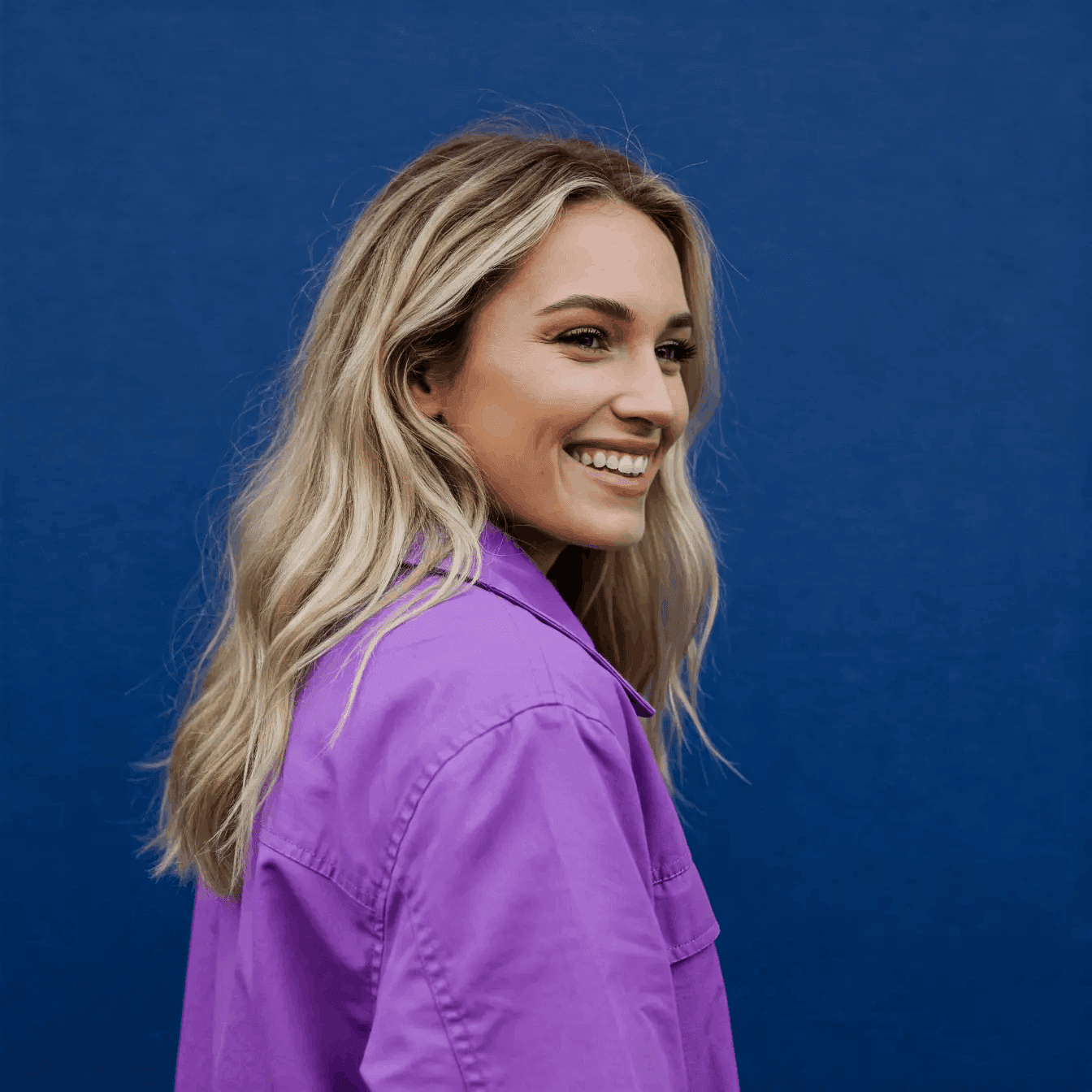 Uśmiechnięta kobieta z blond włosami ubrana w fioletową koszulę, stojąca bokiem na tle granatowej ściany